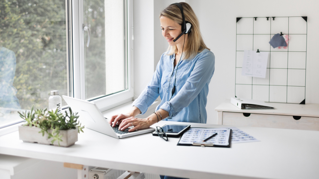 Lady standing at a sit-stand desk in a blue shirt. She is wearing a headset and using a laptop computer for work. 